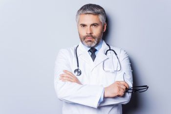 Confident doctor. Mature grey hair doctor looking at camera and keeping arms crossed while standing against grey background